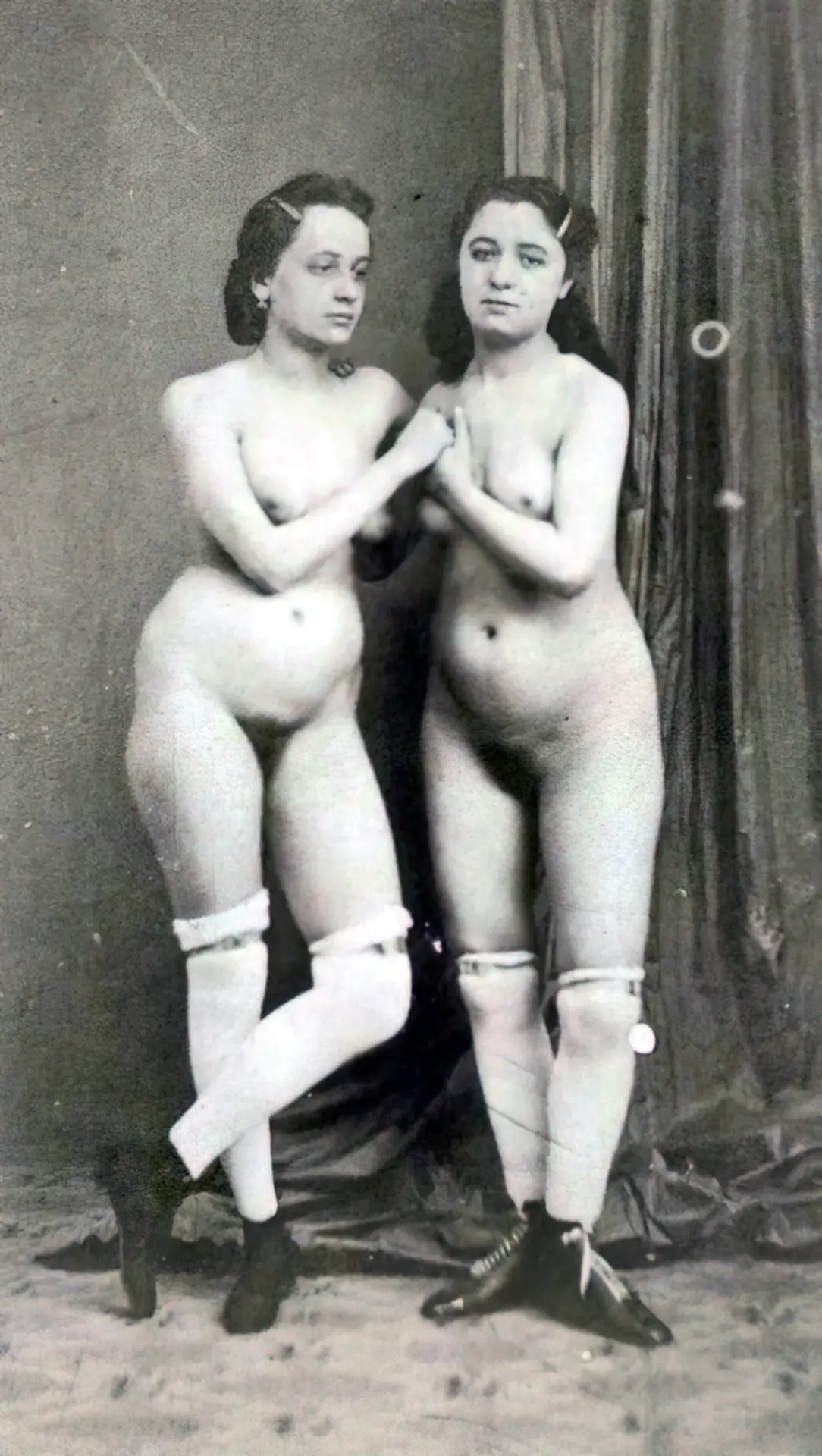 Two sad looking antique women hold each other's hands while posing naked