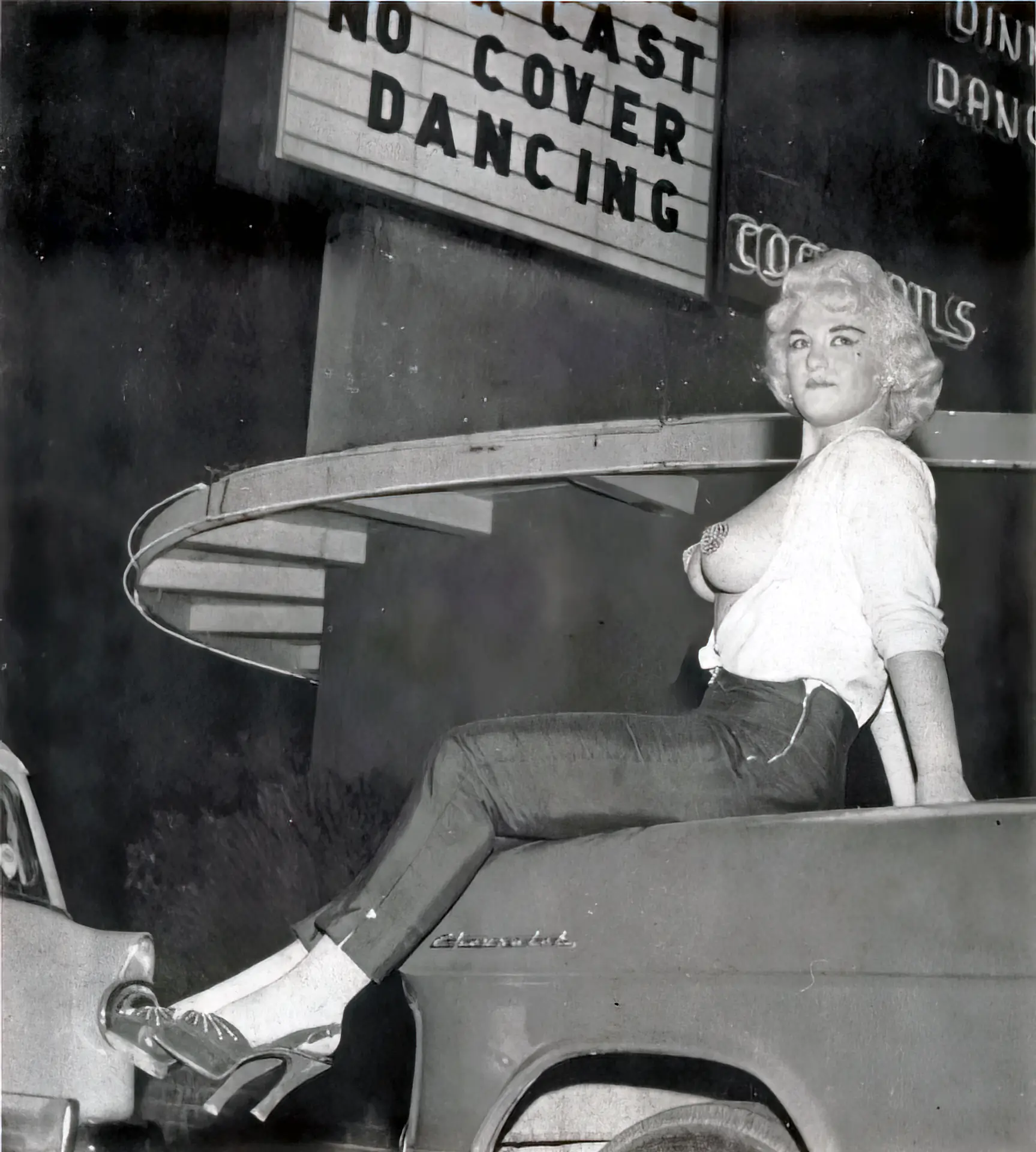 Older burlesque girl sits on a car in an unbuttoned blouse